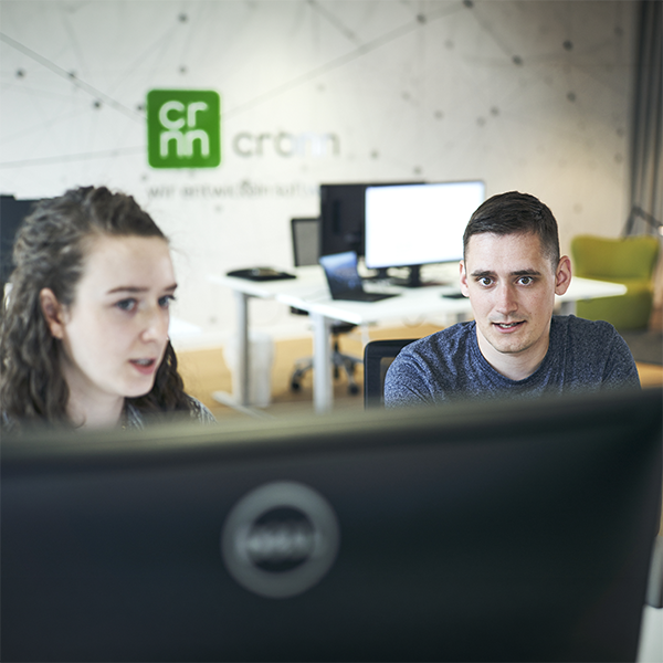 Eine Frau und ein Mann, die auf einen Monitor schauen. Der Mann zeigt auf einen Monitor, darauf sieht man mobile Screens mit einem Prototypen.